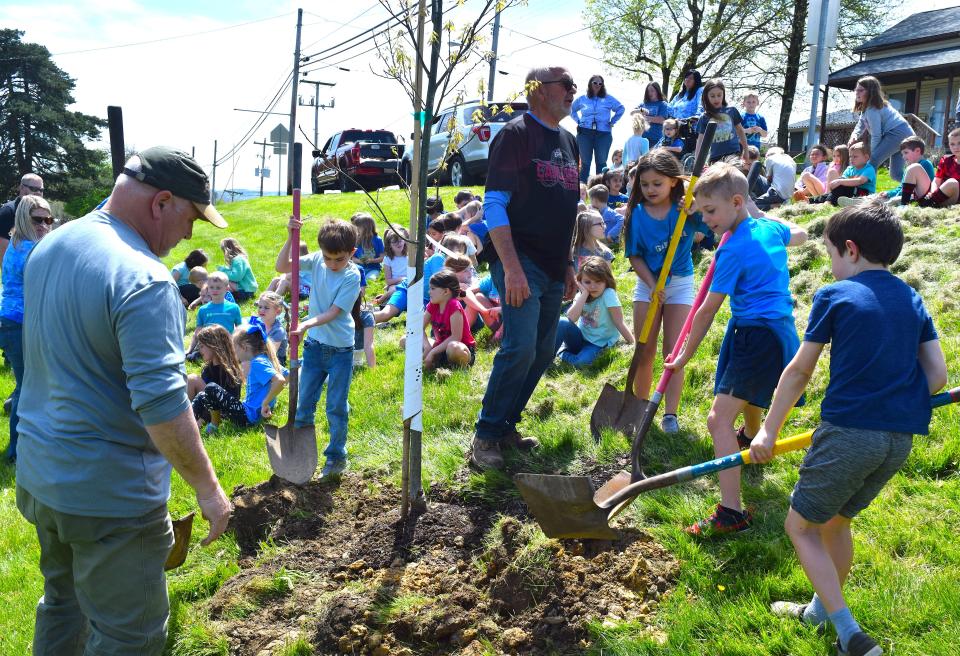 Millersburg Tree City Committee representatives Brent Schrock, left, and Kim Kellogg look on as Millersburg first graders Finlee Morris, Emmy Mellinger, Harrison Rice and Oliver Felder shovel dirt onto the base of a newly planted red oak at Millersburg Elementary School.