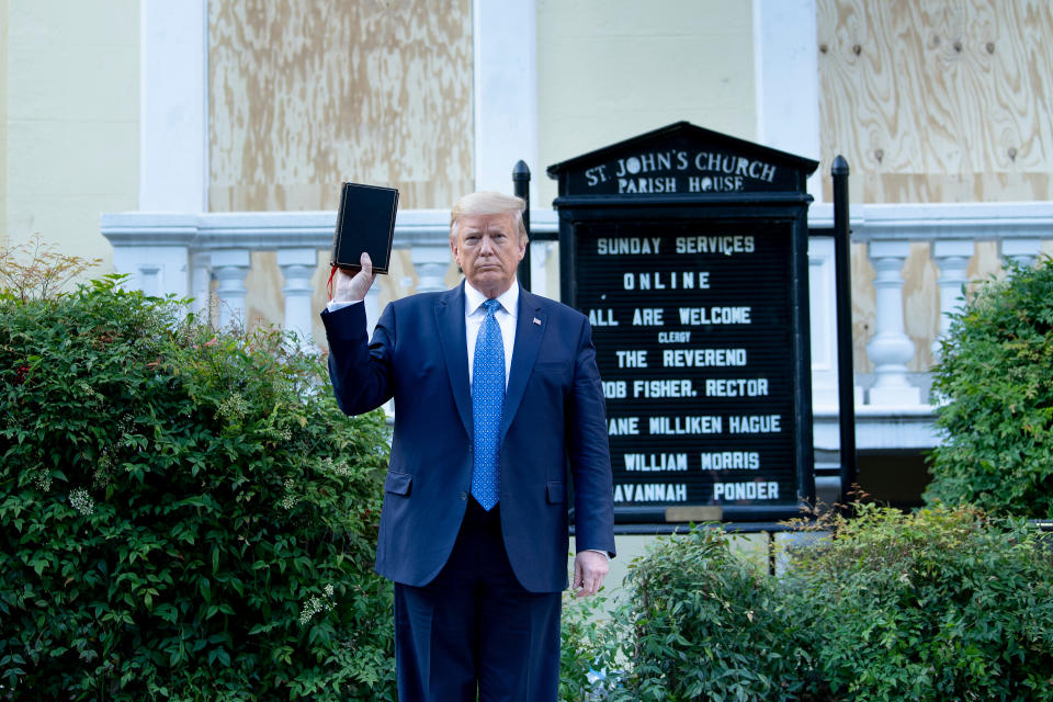Trump holds a Bible aloft in his right hand while standing next to a sign reading St. John's Church Parish House, followed by information on church services.