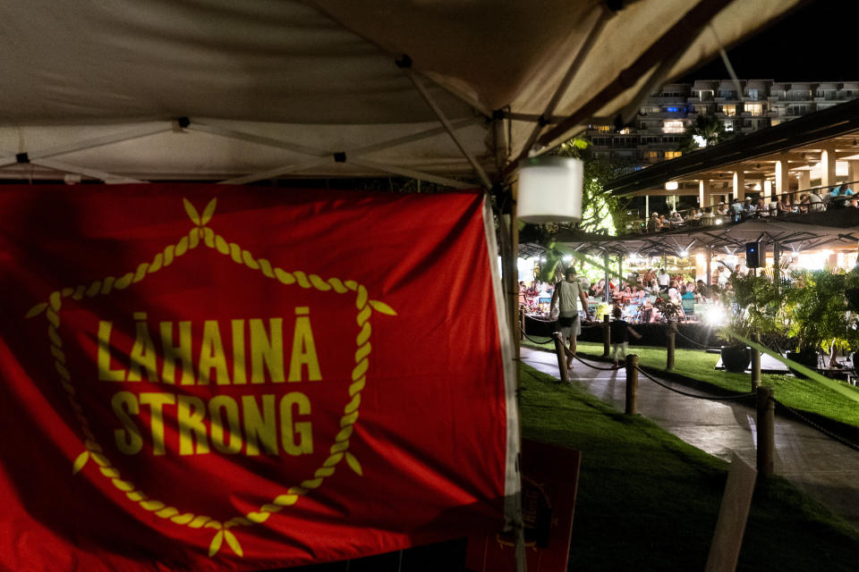 Tourists dine in restaurants at Whalers Village shopping mall in front of a "Lahaina Strong" flag from a housing protest on Kaanapali Beach, Wednesday, Dec. 6, 2023, in Lahaina, Hawaii. A group of survivors is camping on the resort beach to protest and raise awareness for better long-term housing options for those displaced. Residents and survivors still dealing with the aftermath of the August wildfires in Lahaina have mixed feelings as tourists begin to return to the west side of Maui, staying in hotels still housing some displaced residents. (AP Photo/Lindsey Wasson)