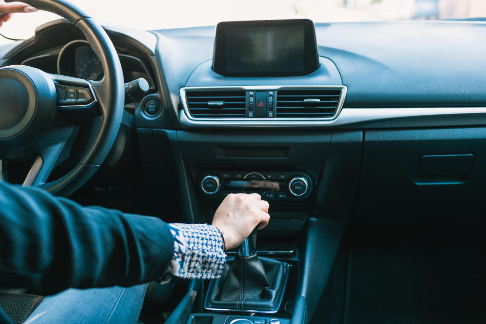 A person wearing a suit is seated in a car, shifting the gear lever with their right hand. The dashboard and steering wheel are visible