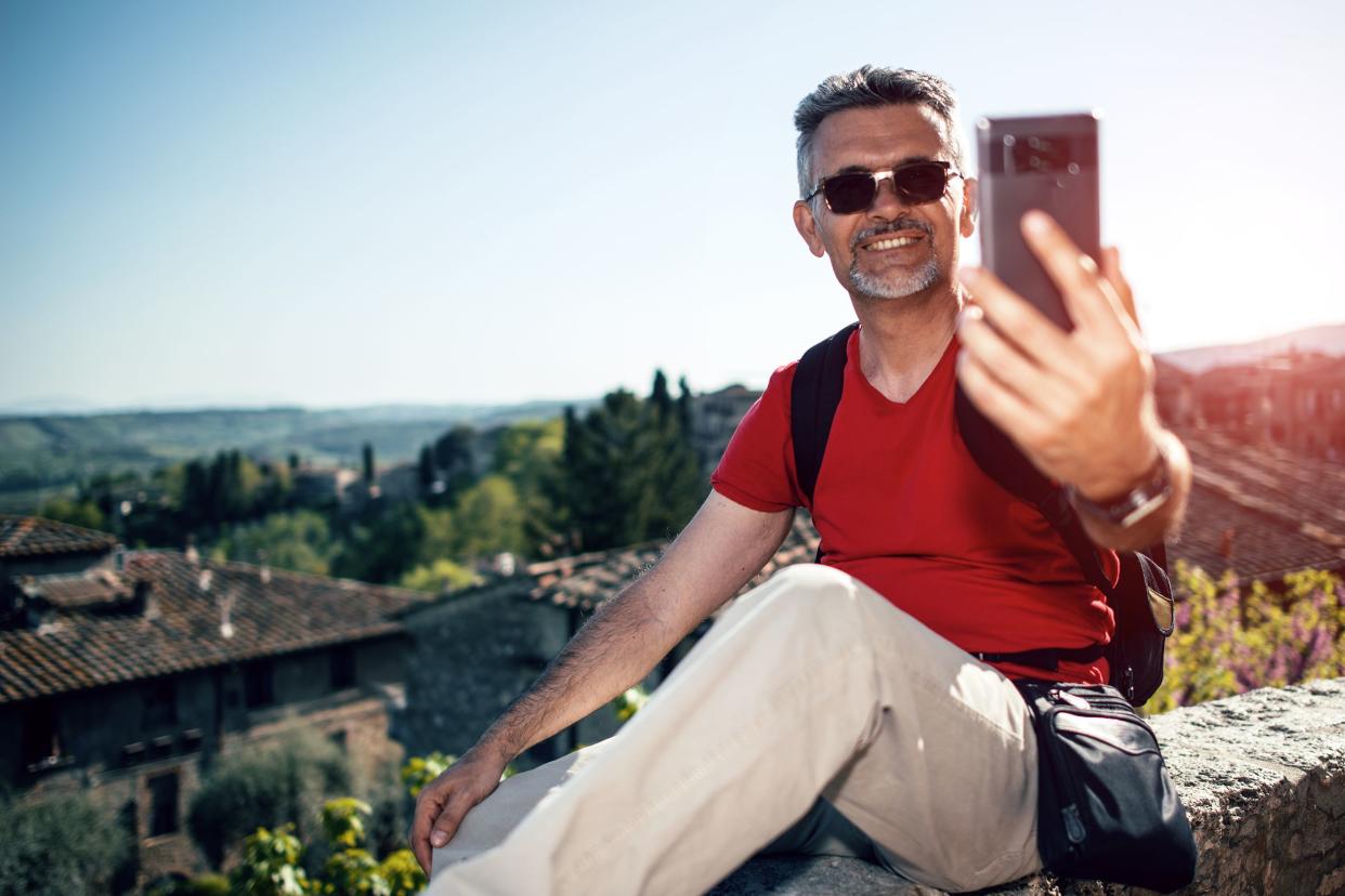 happy handsome senior bearded man smiling while taking selfie picture with mobile phone and wearing eyeglasses outdoors, Florence, Italy