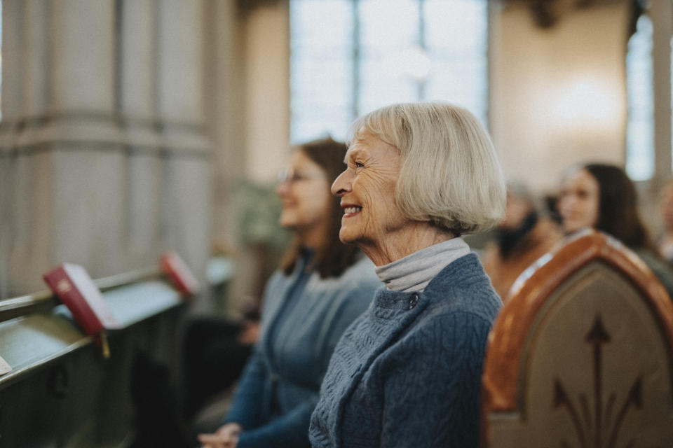 An elderly woman with short gray hair, wearing a textured sweater, smiles while seated in a pew inside a church, with other people around her