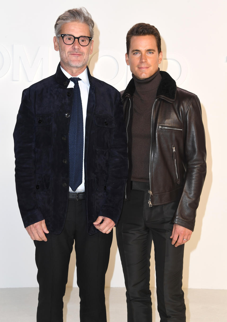 Two men pose together, one wearing a velvet blazer and glasses, the other wearing a leather jacket and turtleneck