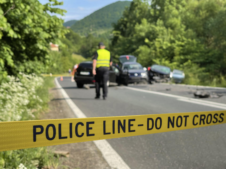 Police line tape blocks a road as an officer in a yellow vest investigates a multi-car accident. Several vehicles are damaged in the background