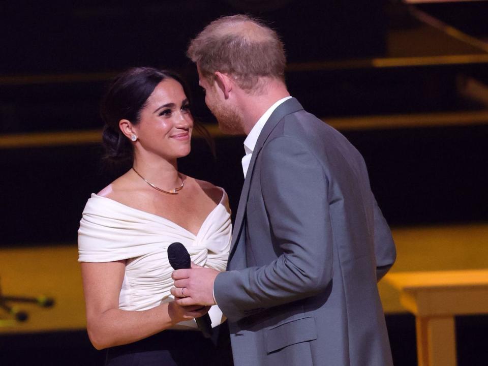 Meghan in a white off the shoulder blouse smiling at Harry who has his back to the camera and is holding a microphone, leaning in to give Meghan a kiss.
