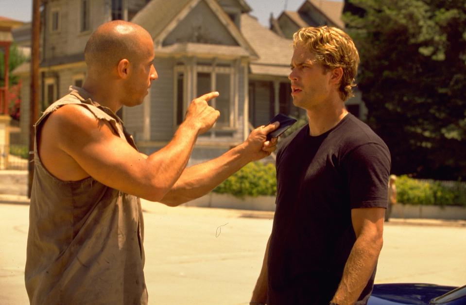 in a scene, Vin Diesel points at Paul Walker while holding a phone, with both standing outside in front of houses