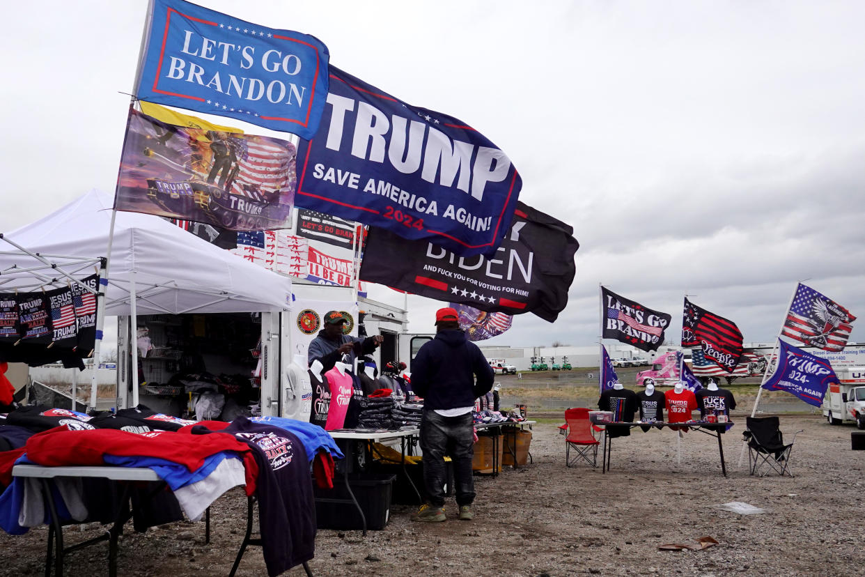 A vendor talks to a customer by his stacked blue and red merchandise at a booth with flags reading: Let's Go Brandon and Trump, Save America Again.!