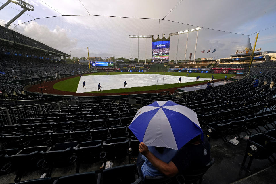 Fans wait under an umbrella during a rain delay in the second inning of a baseball game between the Kansas City Royals and the Milwaukee Brewers Tuesday, May 18, 2021, in Kansas City, Mo. (AP Photo/Charlie Riedel)