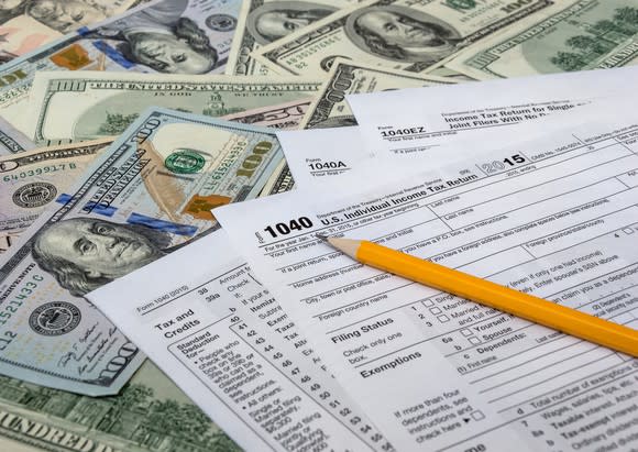 Tax forms on top of a spread-out pile of U.S. currency, with a pencil on top of the pile.