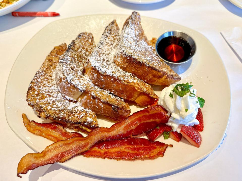 Four half-pieces of French toast with a cup of syrup, whipped cream with chopped-up mint, and bacon strips on the side. The French toast is sprinkled with powdered sugar