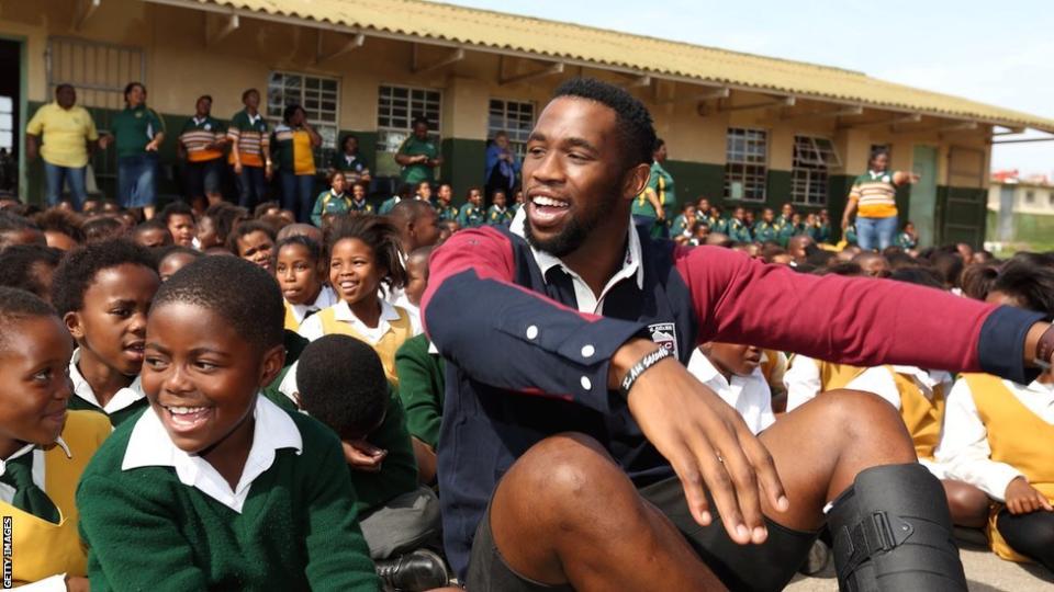 South Africa captain Siya Kolisi sits on the floor with a large group of young schoolchildren sitting around and behind him in front of a school building