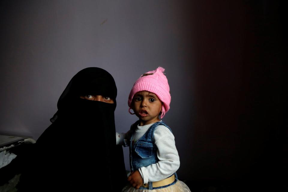 Woman in black burqa holds a very skinny child wearing a pink hat, in a hospital setting