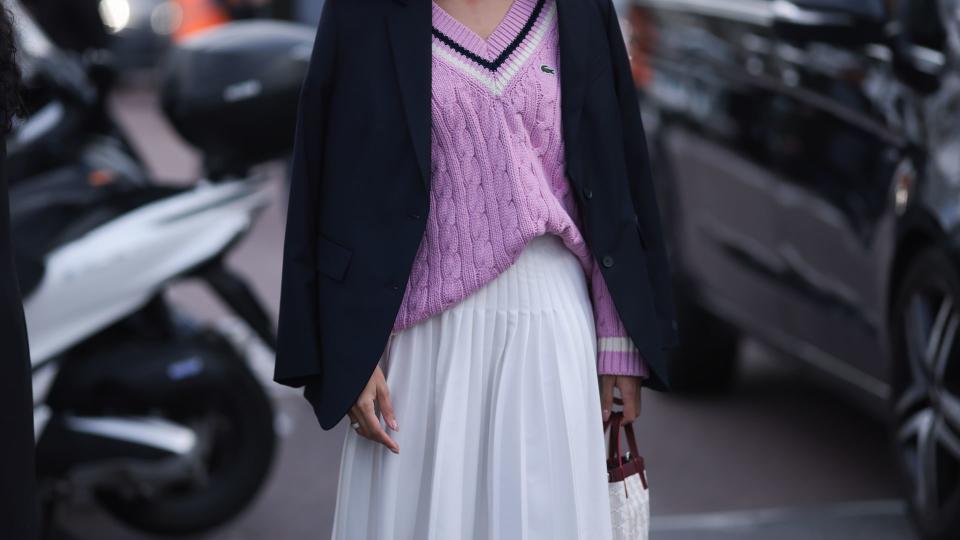 A Guest seen wearing a black blazer jacket, purple Lacoste pullover, white skirt, socks, green sneaker, black shades and white bag outside Lacoste Show during the Womenswear Fall/Winter 2024/2025