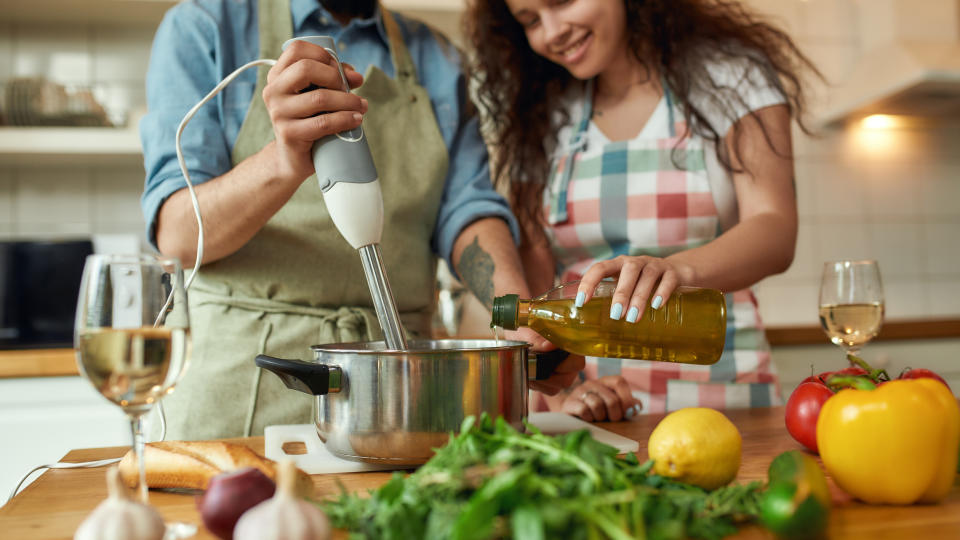 Cropped shot of man, chef cook using hand blender while preparing a meal. Young woman, girlfriend in apron pouring olive oil in the pot, helping him in the kitchen. Cooking at home. Web Banner