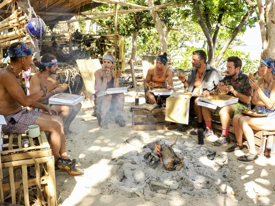 Survivor players sitting at camp and eating pizza out of boxes, with the forest in the background