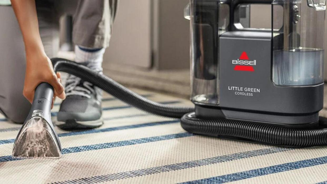  An olive-skinned female using the Bissell Little Green Cordless Portable Carpet Cleaner on cream and blue-striped area mat. 