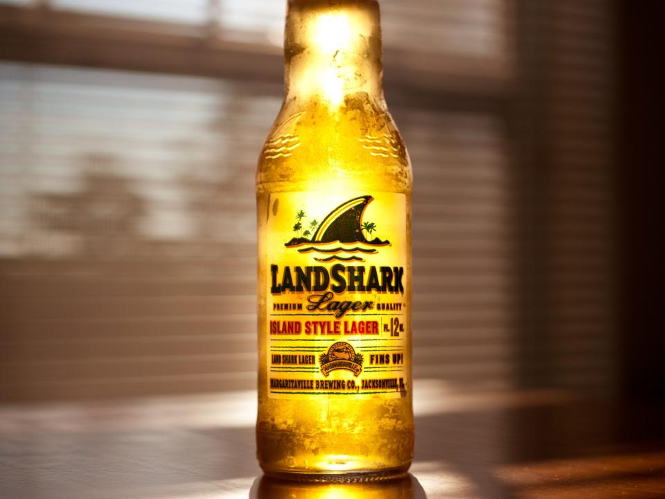 Front view of an open bottle of Landshark Island Style Lager/Beer. The bottle features a clear label with a shark tail and styled text. The bottle sits alone on a table, back lit by afternoon sun. A reflection and shadow are cast in front of the bottle. A large window can be seen out of focus in the background.