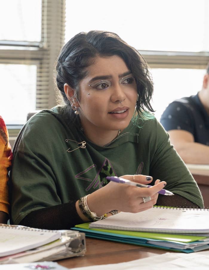 Woman in green top with safety pins, looking concerned, holding pen over notebook in a classroom setting