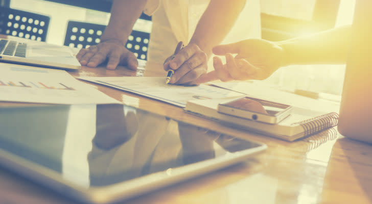 Two people take notes on paper in a brightly-lit room with a laptop, a tablet, and a phone all on the same table.