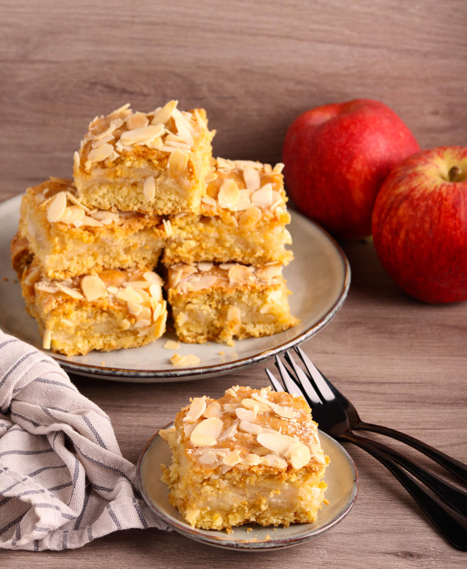 A plate of almond-topped cake squares is stacked beside two apples and a smaller plate with a cake square. Two forks and a napkin are also in the image