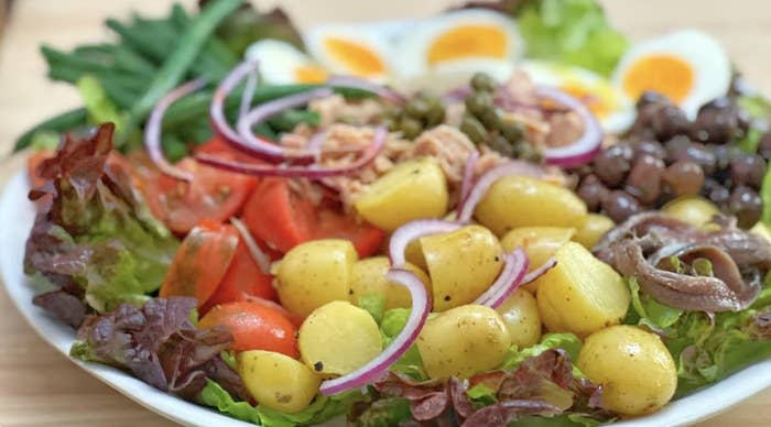 A plate of salad with potatoes, tomatoes, green beans, lettuce, red onions, eggs, anchovies, olives, and capers