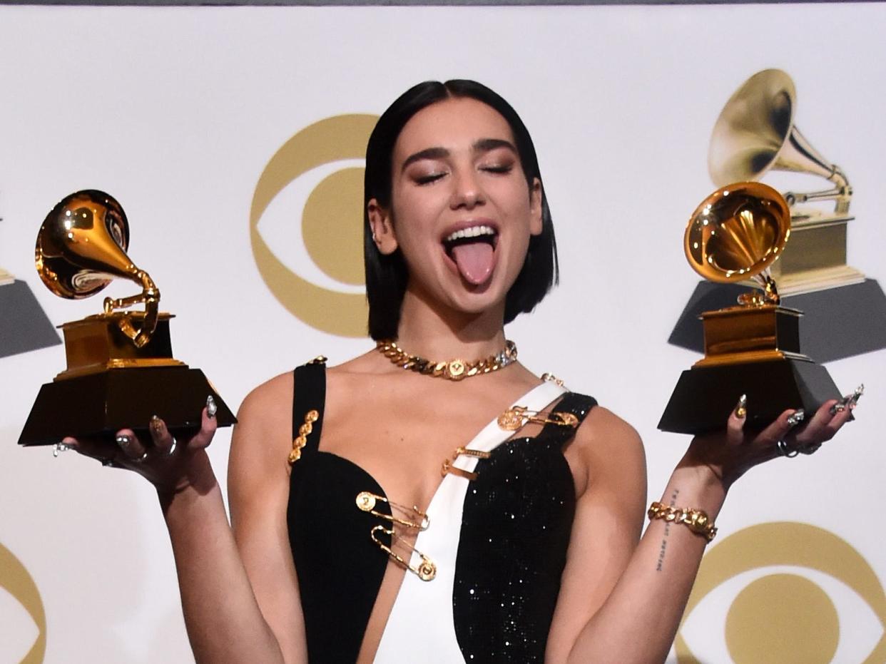 Dua Lipa holds two grammys and stands in black and white dress while sticking her tongue out.