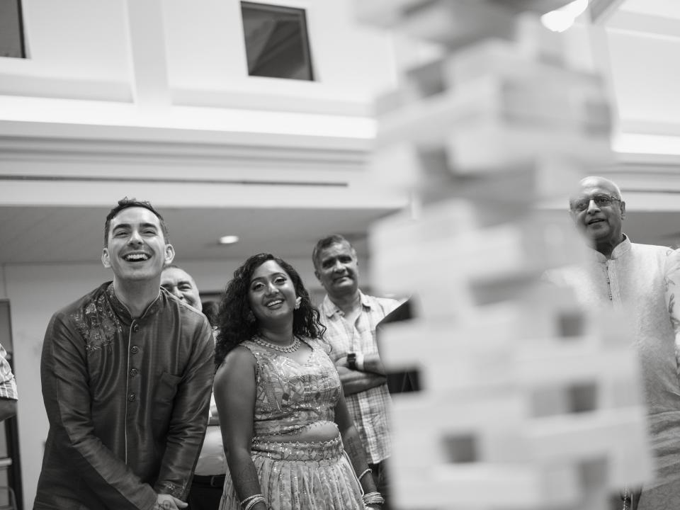 The couple are standing to the left of the shot in traditional Indian clothing, and are smiling with their guests at a Jenga tower, shown out-of-focus to the right of the shot