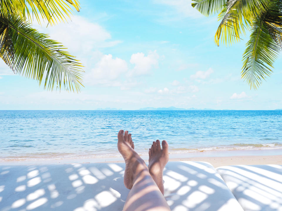 Woman feet on sunbath chair with coconut leaves shadow over summer beach with blue sky and white clouds landscape background.