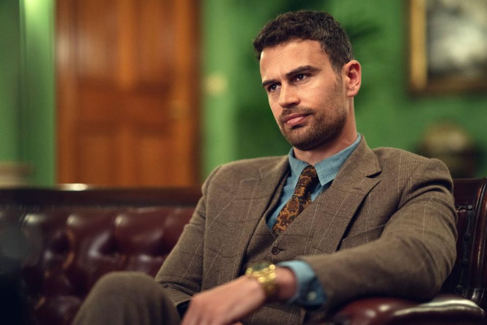 Theo James sits on a leather couch, wearing a stylish suit and patterned tie