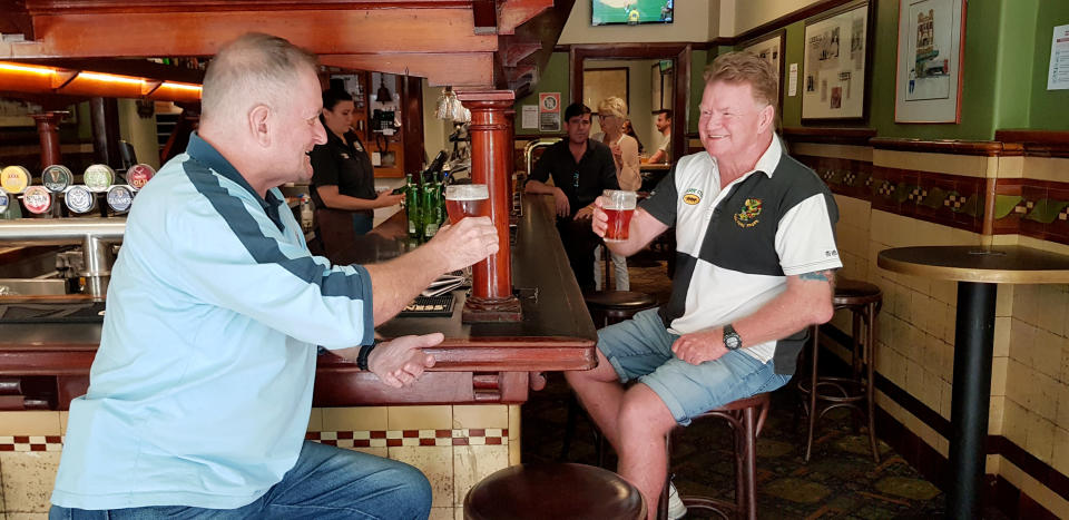 Two friends sit at a bar inside a pub and hold up their beers while looking at each other. 