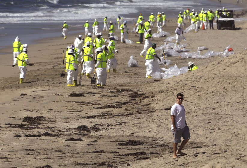 HUNTINGTON BEACH, CA - OCTOBER 5, 2021 - - A beachcomber walks past workers who continue to clean the beach from an oil spill earlier this week at Huntington Beach on Saturday, October 5, 2021. (Genaro Molina / Los Angeles Times)