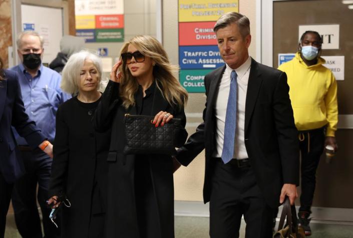 A woman wearing sunglasses is led through a building by a man in a suit.