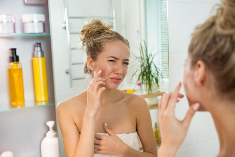 Young beautiful woman touching skin in bathroom. Unhappy girl standing in towel, looking in the mirror, checking dry irritated skin. Morning skincare routine.