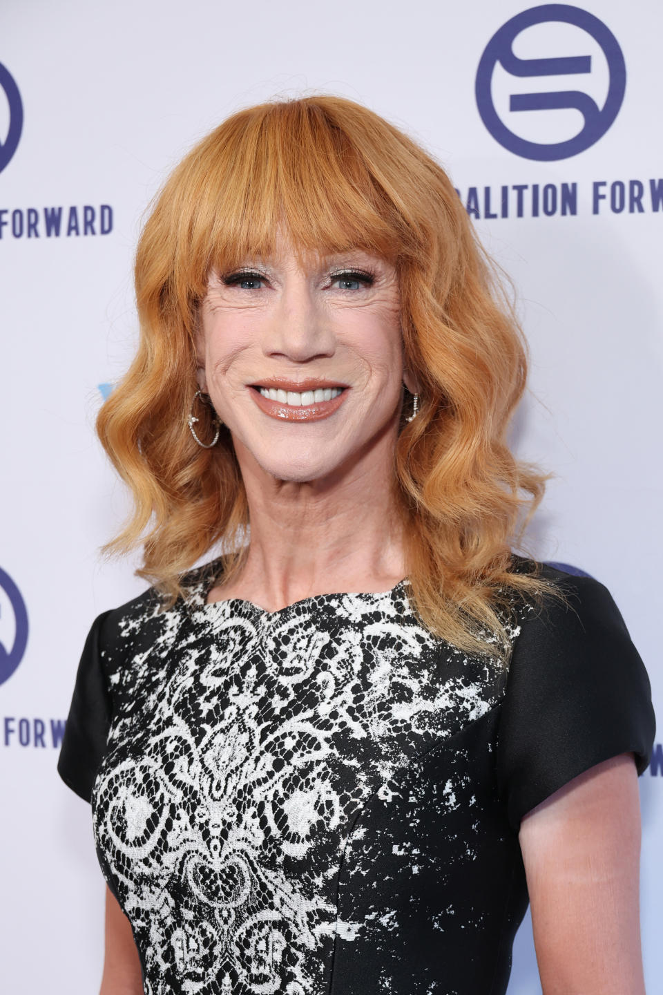 Kathy Griffin smiles at an event, wearing a black dress with intricate white patterns on the front