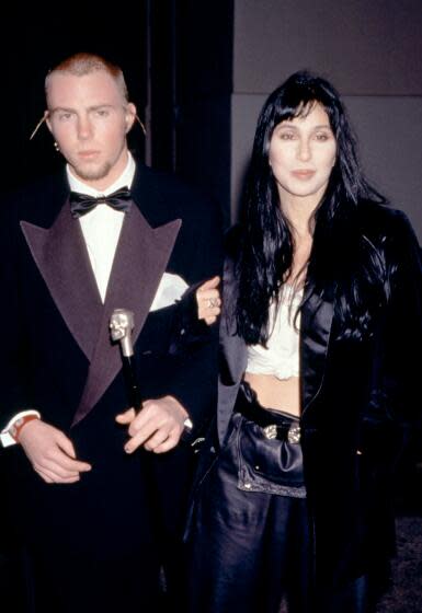 Elijah Blue Allman wears a tuxedo and is linking arms with his mother Cher, wearing a long black coat and white crop top