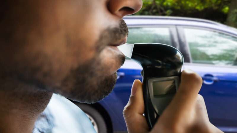 A male motorist blows into a Breathalyzer while sitting in the front seat of his car.