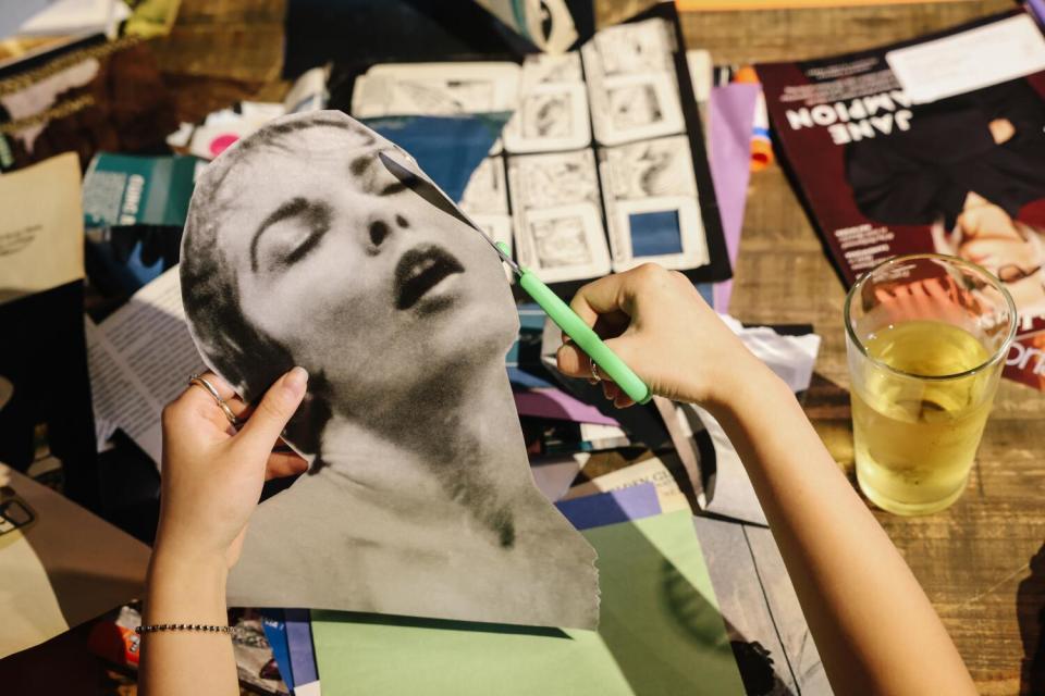 A hand using scissors to cut out a black-and-white photo of a woman's face, with a glass of wine nearby.