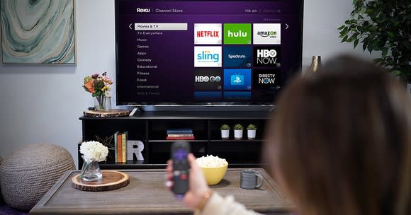 A person pointing a Roku remote at a TV displaying the Roku homescreen.