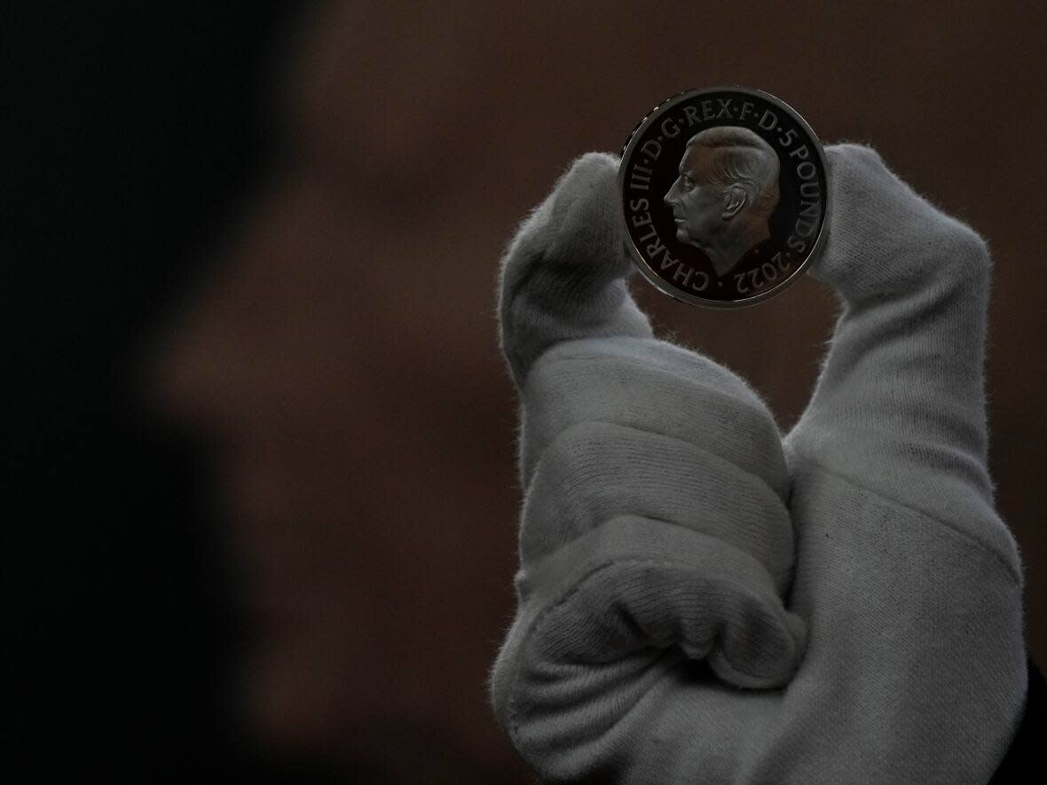 A staff member with the U.K.'s Royal Mint holds up a commemorative five-pound coin bearing the official coinage portrait of King Charles III. Canada will soon follow suit, producing new coins and banknotes bearing an original image of the new monarch.  (Alastair Grant/The Associated Press - image credit)