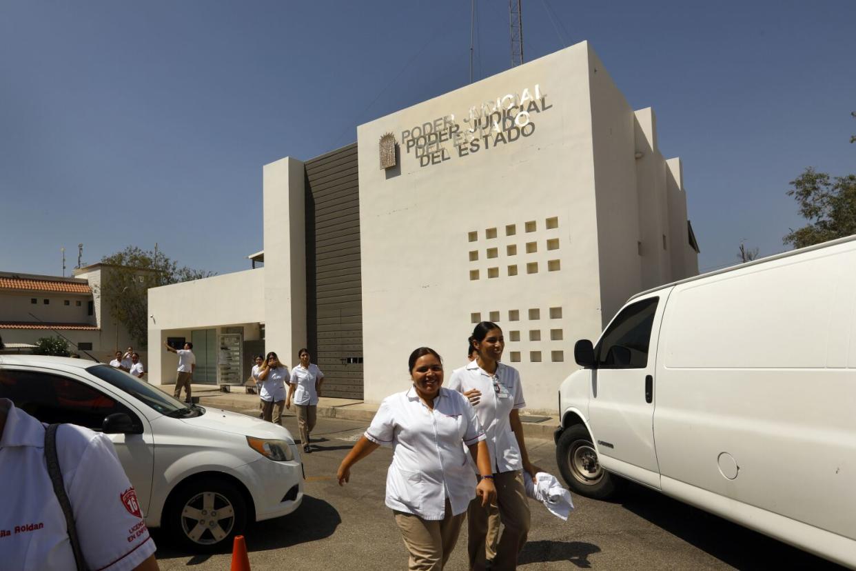 Two women in a white uniform and khaki pants walk near a beige-colored building and cars