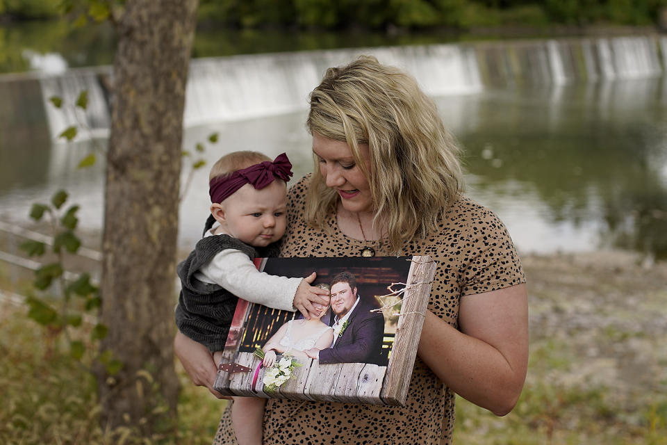 Aubrea Baker displays her wedding photo as she and her 7-month-old daughter Haylen visit one of her late husband's favorite fishing spots Saturday, Oct. 2, 2021, in Burlington, Kan. Her husband, Danny Baker, was among the 700,000 U.S. victims of COVID-19, dying on Sept. 14, after testing positive in July. (AP Photo/Charlie Riedel)