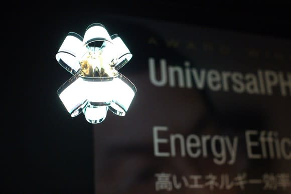 A futuristic lighting fixture featuring flexible OLED panels hangs in front of a black trade show banner.