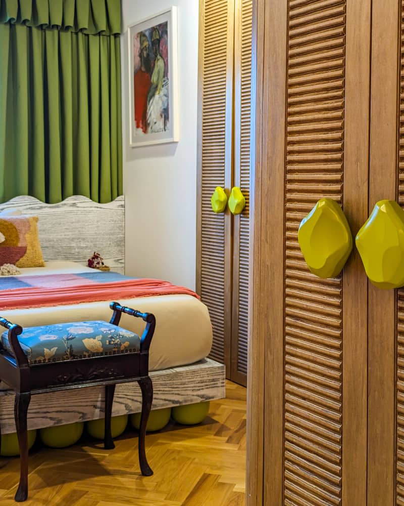 Bedroom with tree branch wallpaper wall, green curtains, parquet wood floor, wall of wood closets, and pops of yellow and orange