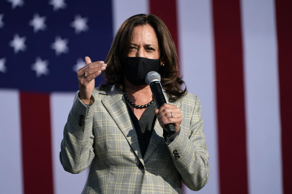 Sen. Kamala Harris speaks into a microphone wearing a black face mask and standing in front of an American flag