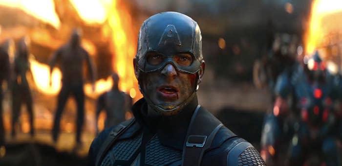 Chris Evans, as Captain America, stands determined in battle gear with a scarred face, surrounded by allies in the midst of an intense fight scene