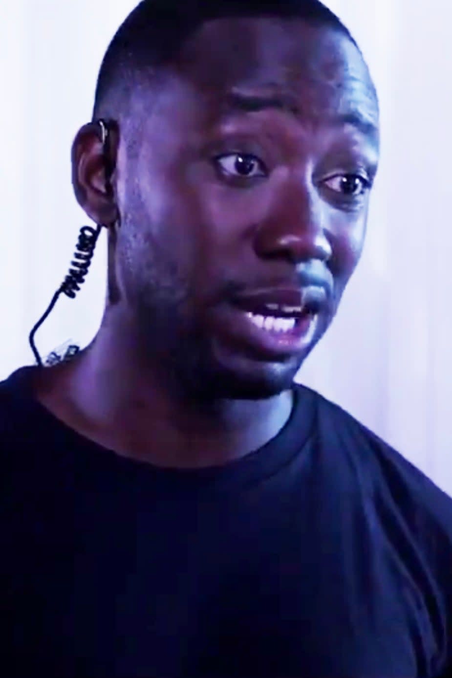 Lamorne Morris with a concerned expression, wearing a black shirt and an earpiece in a TV scene