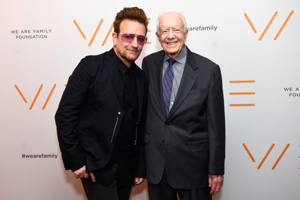 bono, wearing sunglasses and a black coat and shirt, standing next to a smiling jimmy carter, wearing a great suit and blue and red striped tie