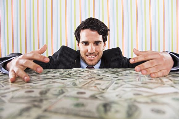 A smiling businessman, eyes wide open, admiring a messy pile of cash on a table with his arms around the dollar bills as if trying to embrace them.