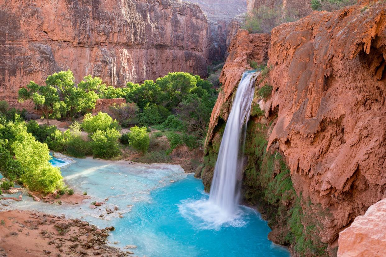 Havasupai Falls, Grand Canyon National Park, Arizona, waterfalls cascading into blue swimming hole surrounded by terracotta colored rocks and some green trees and plants to the left