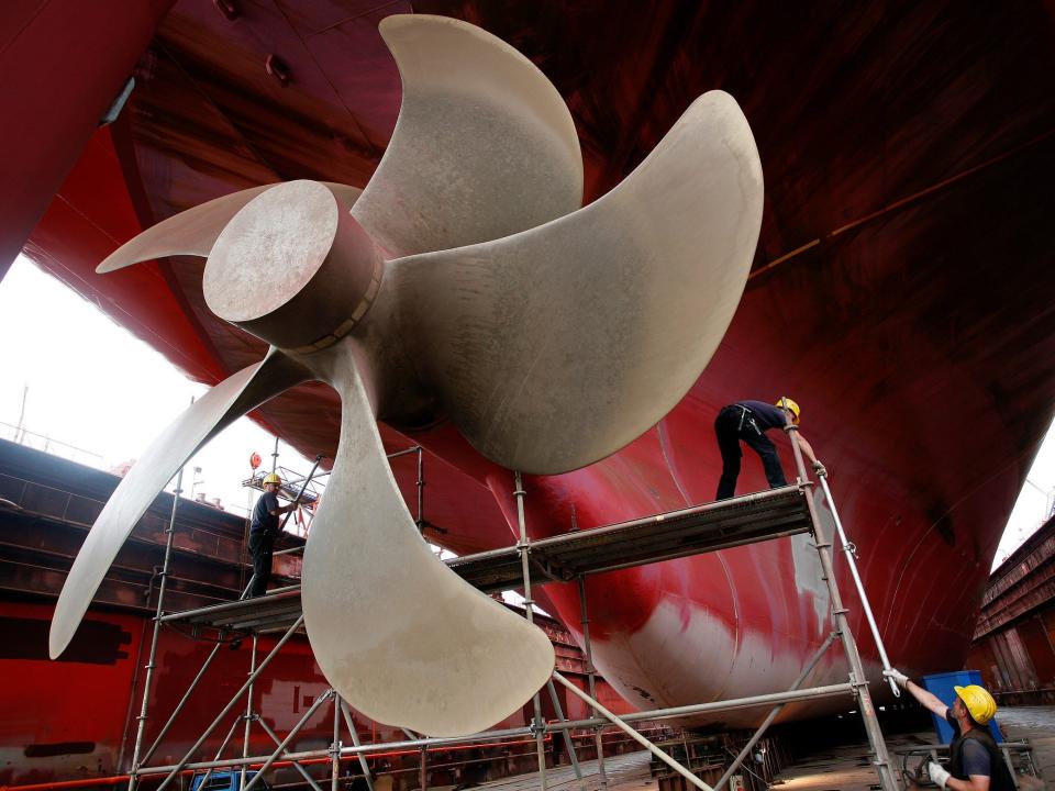 two workers in yellow hardhats pass a pole up scaffolding beneath a huge ship propeller with spiraling blades larger than the people, attached to a giant red ship